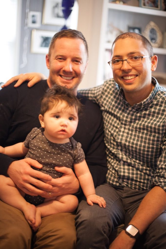 Derek and Jonathan with large smiles holding their daughter in their home